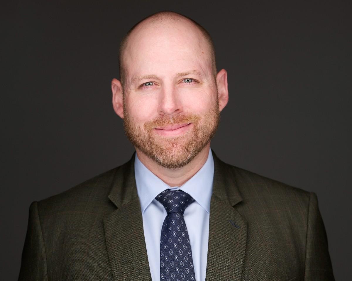 Bald man with a short beard wearing a suit and patterned tie, smiling slightly, photographed against a plain dark background.