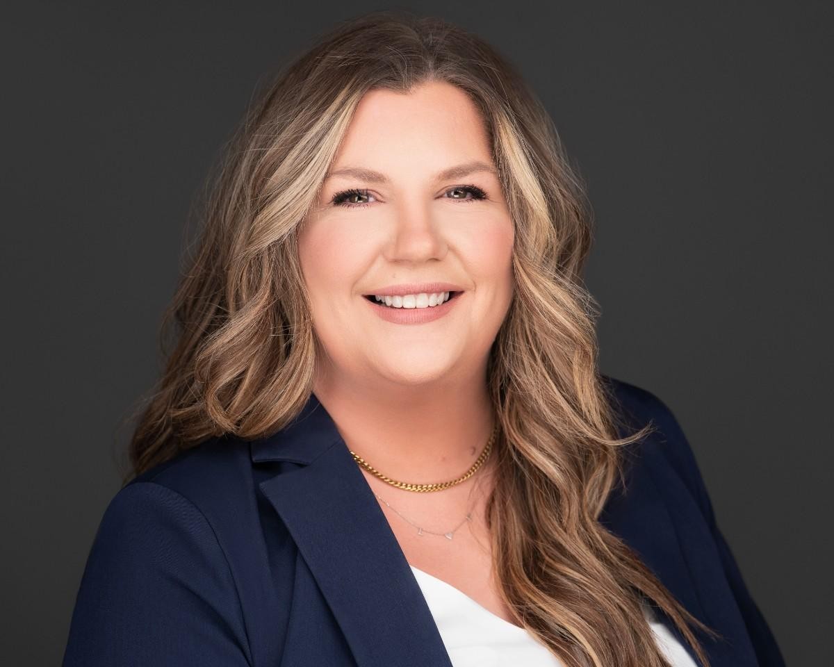 A woman with long wavy hair wearing a navy blazer, white top, and gold necklaces, smiling in front of a dark background.