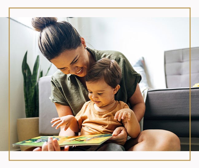 Woman sitting on the floor reading a picture book with a young child in a living room with a plant and sofa in the background.