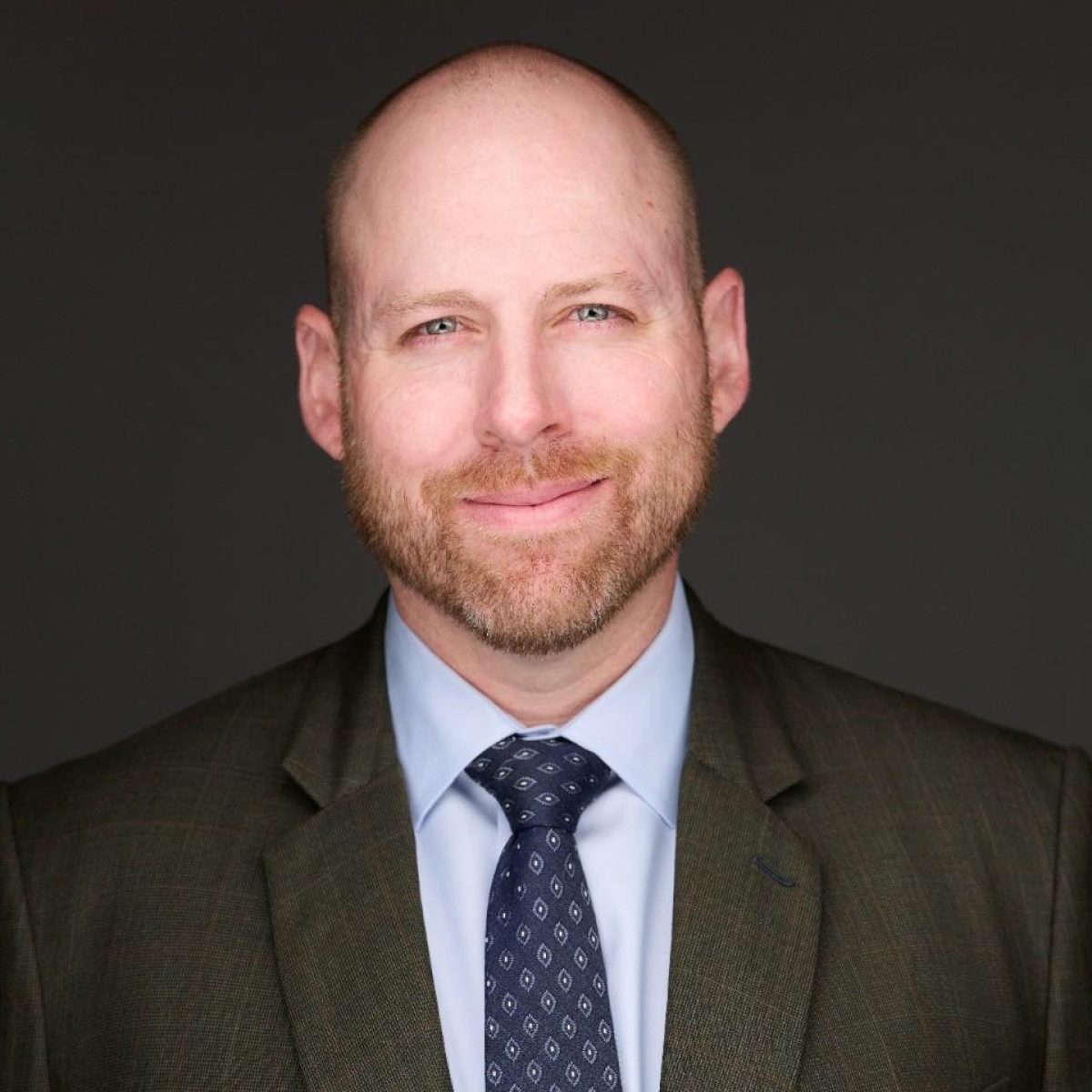 Bald man with a short beard wearing a suit and patterned tie, smiling slightly, photographed against a plain dark background.