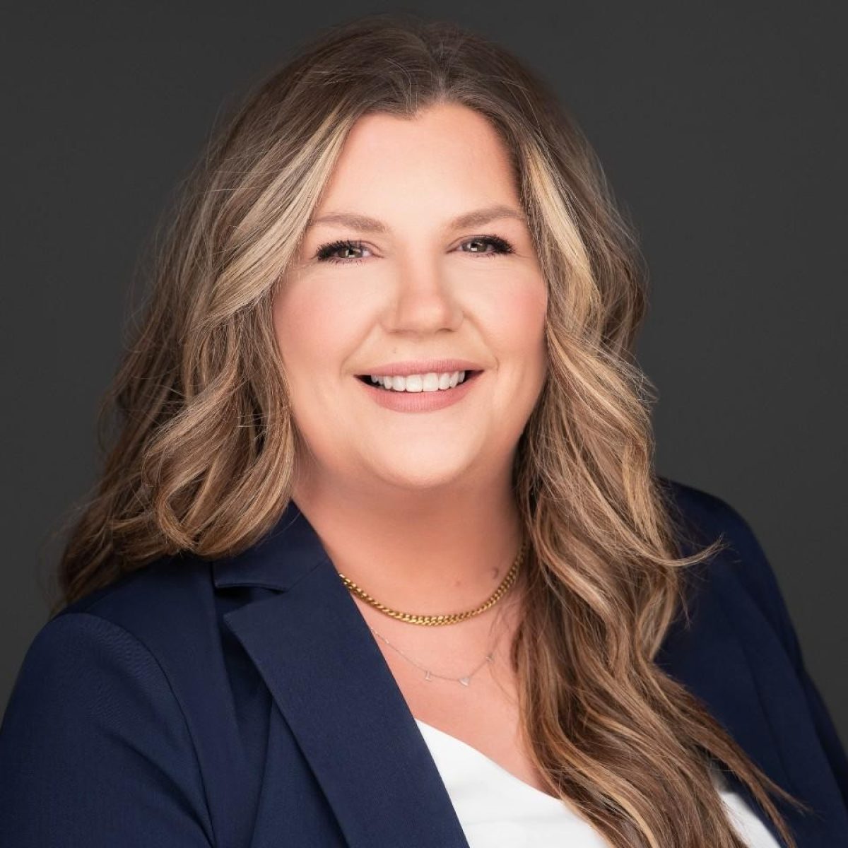 A woman with long wavy hair wearing a navy blazer, white top, and gold necklaces, smiling in front of a dark background.