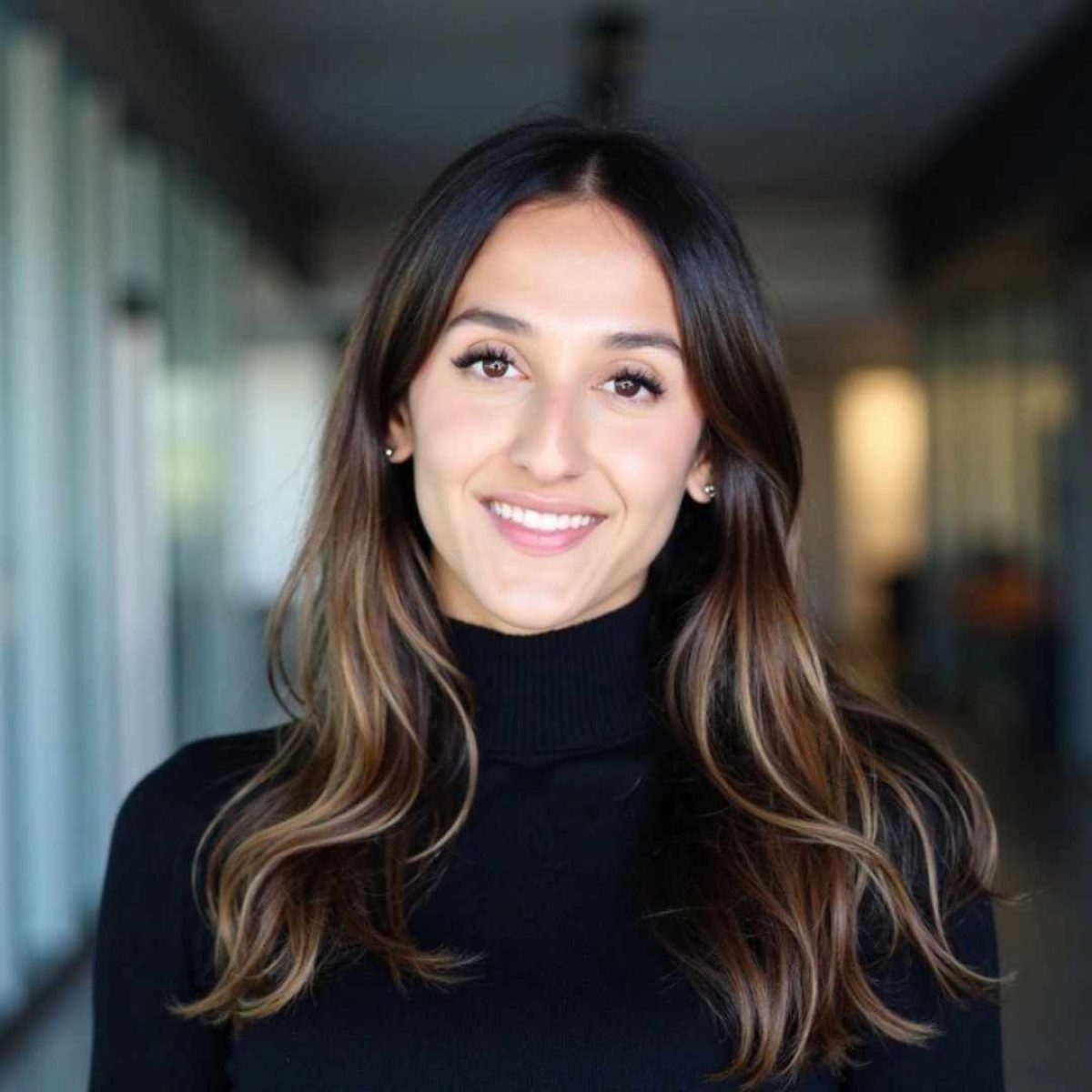 A woman with long brown hair, wearing a black turtleneck, smiles at the camera in a well-lit hallway with glass walls.